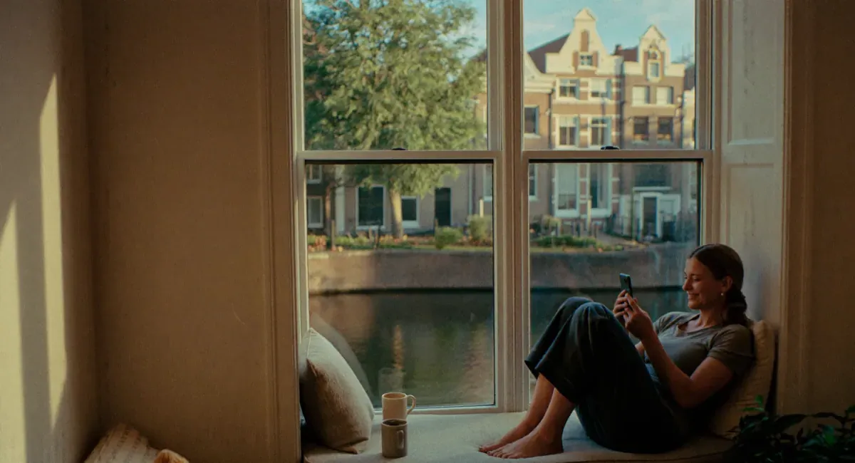 A woman sits smiling on the window seat of a canal house, looking at her phone.